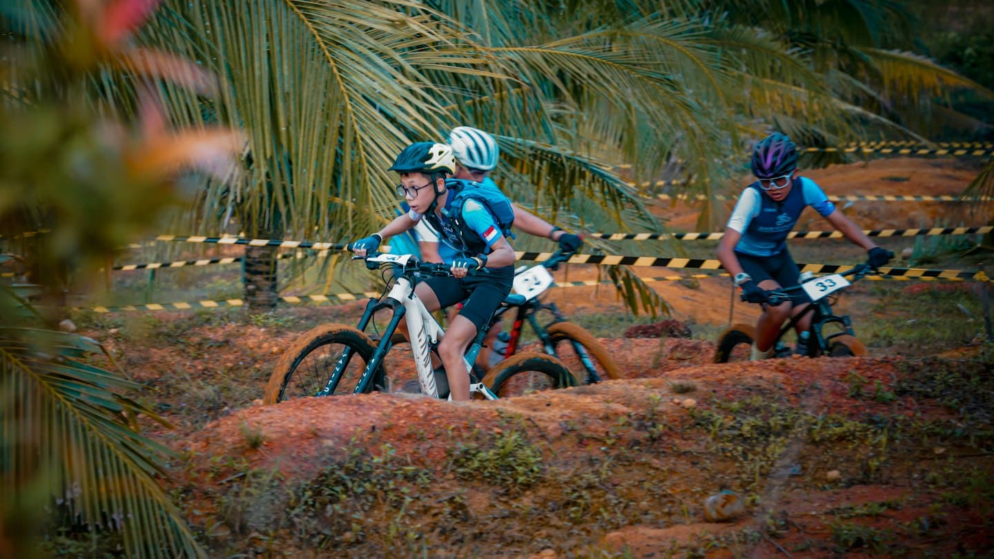 Drei Mountainbiker auf einem schlammigen Pfad nahe Palmen.