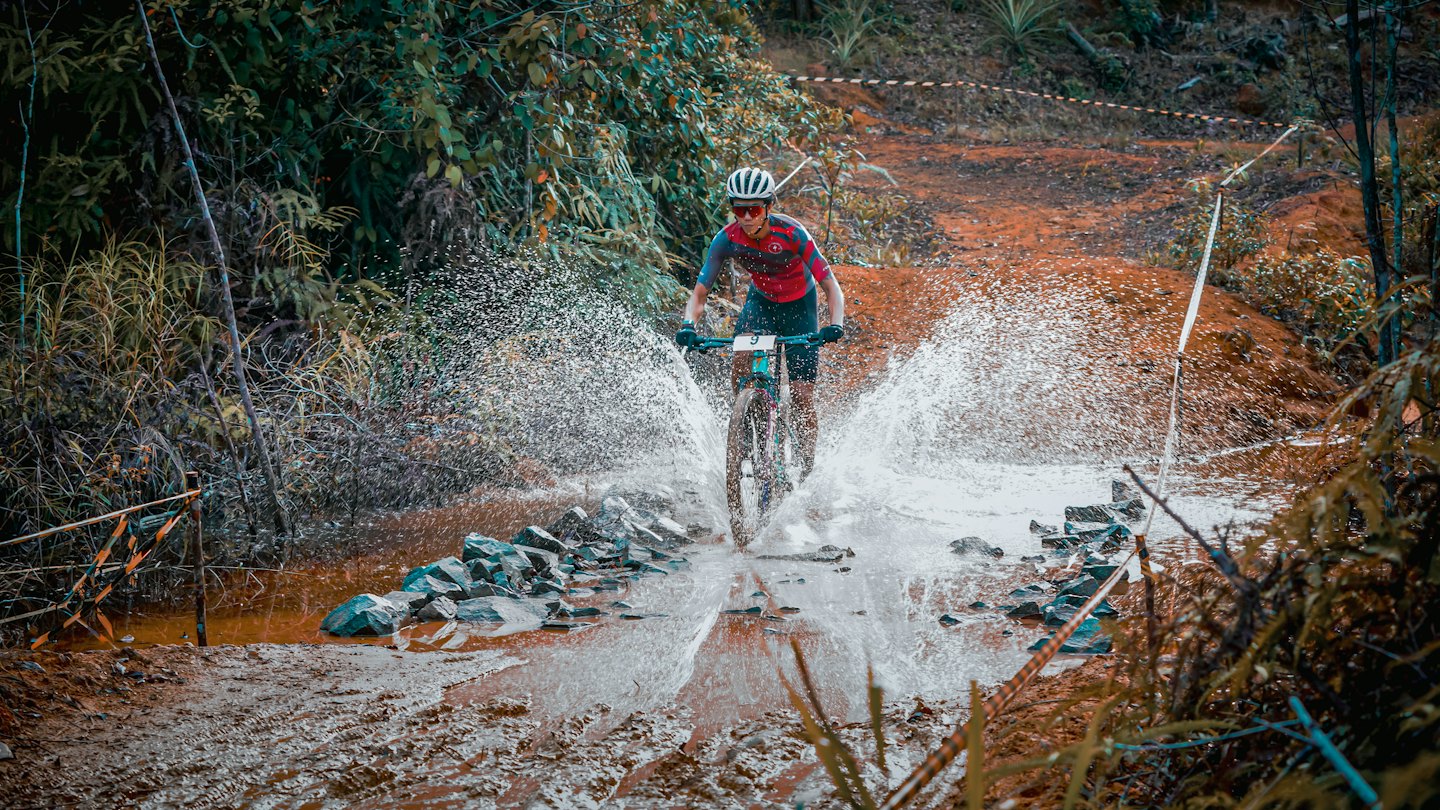 Radfahrer im roten Trikot durchquert spritzend einen schlammigen Bach.